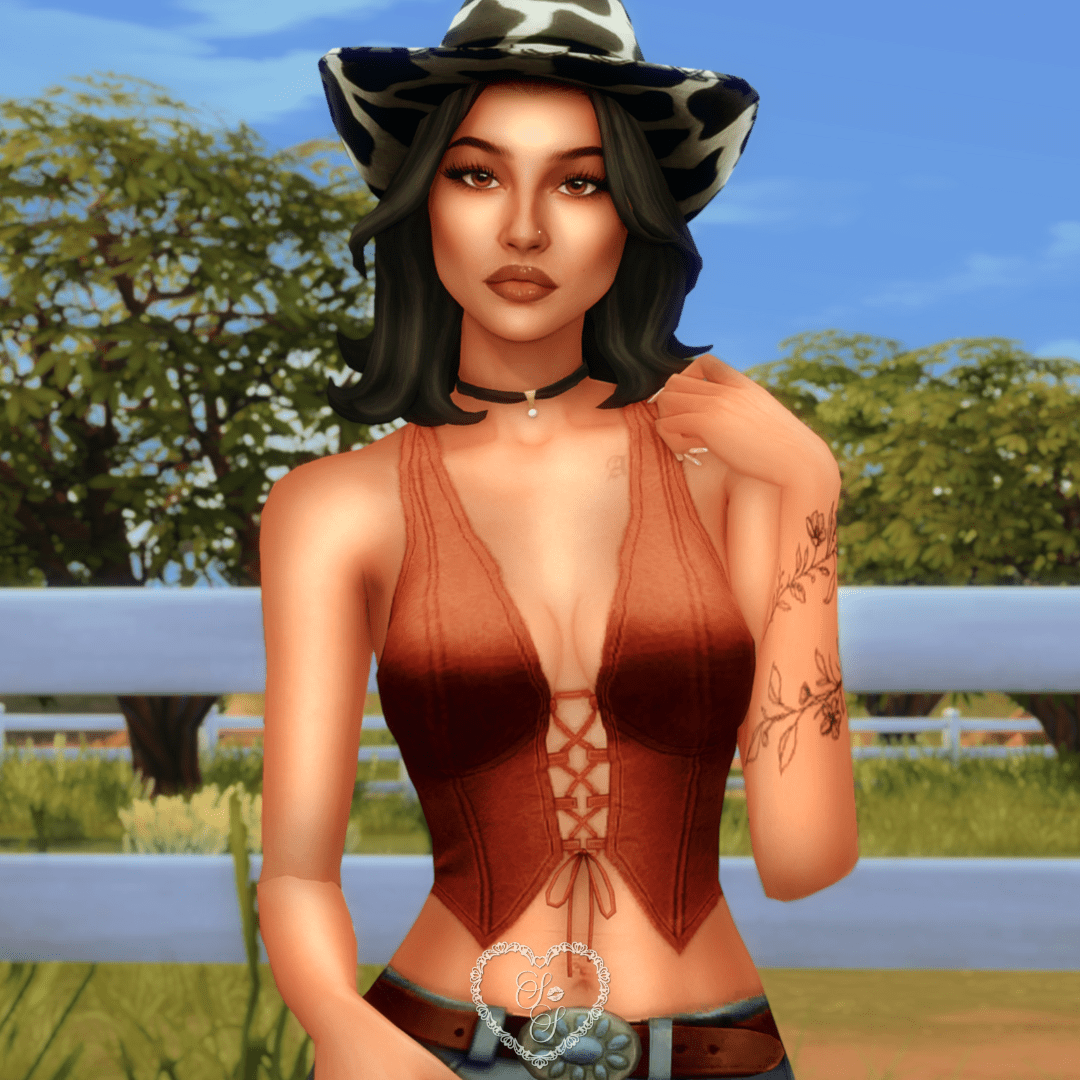 Close-up of Camila showing off her dark wavy hair, cow-print hat, rust-toned Bonnie top, and soft glam makeup in a western farm setting.
